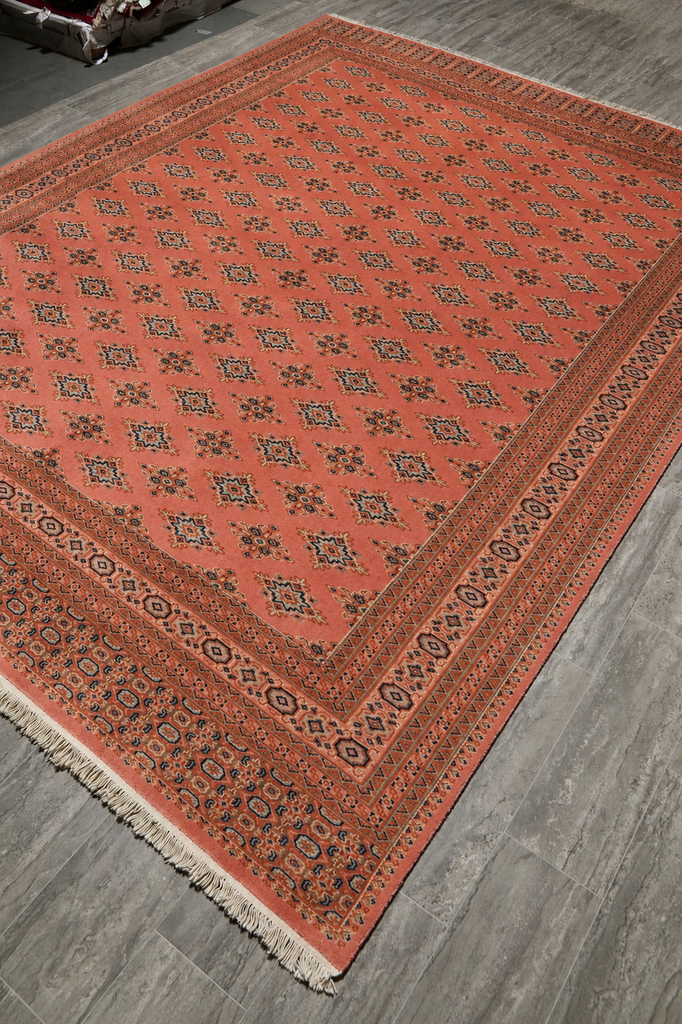 A rectangular area rug with a terracotta orange-red field and intricate geometric borders, displayed on a gray wood-plank floor.