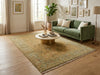 Load image into Gallery viewer, A large traditional-style area rug in muted beige, olive green, and cream tones with fringe, shown in a styled living room setting with a green sofa, round coffee table, wood floors, and framed wall art.