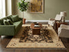 Load image into Gallery viewer, A large traditional-style area rug in beige, brown, and black is styled in a living room setting with a green sofa, wooden coffee table, beige armchairs, and a potted plant.