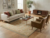 Load image into Gallery viewer, A styled living room scene featuring a beige upholstered sofa with rust and sage green pillows, two brown armchairs, a wooden coffee table, a patterned area rug, and a dark wood media console against a warm beige wall.
