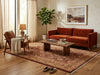 Load image into Gallery viewer, A styled living room featuring a rust-orange velvet sofa as the main product, placed on a large patterned area rug with a wooden coffee table, armchair, lamp, and potted plant in a warm neutral interior.