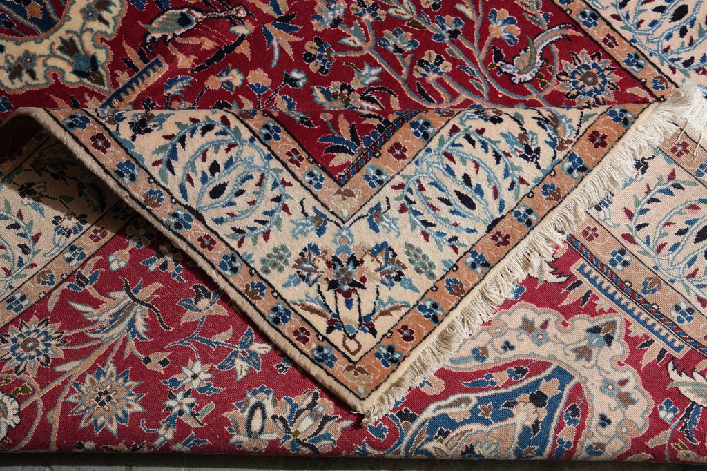 A close-up of a traditional patterned rug with a deep red field, beige borders, and blue floral motifs, showing folded sections and fringe.