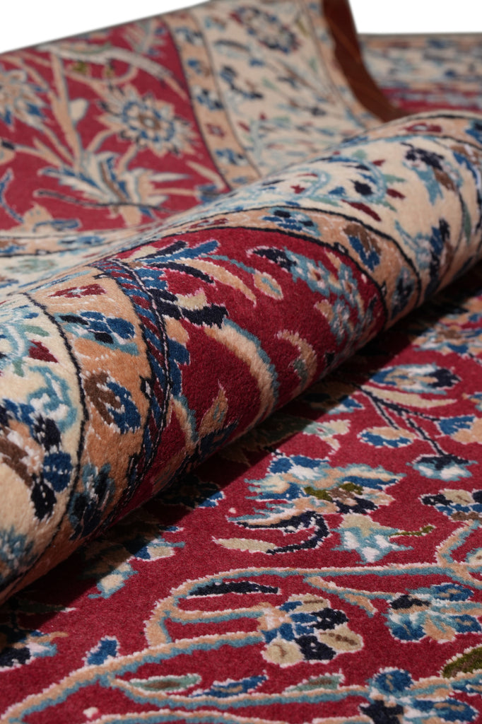 A close-up of a rolled traditional patterned rug showing its red, beige, and blue floral design against a matching flat section of the rug.