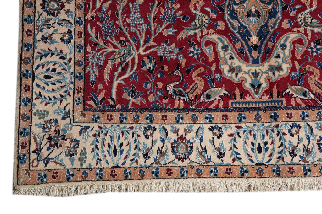 A close-up of an ornate patterned rug corner showing a deep red field with cream, blue, and beige floral and bird motifs, plus a decorative border and fringe.