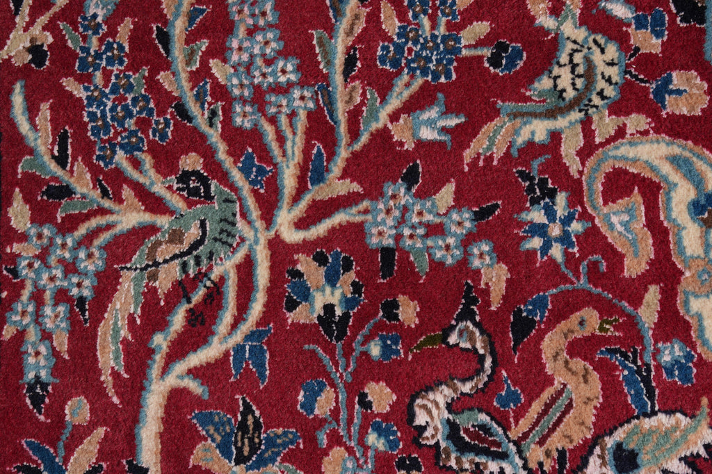 A close-up of a traditional patterned rug featuring a deep red background with beige branches, blue floral clusters, and stylized birds in muted green, blue, and beige tones.