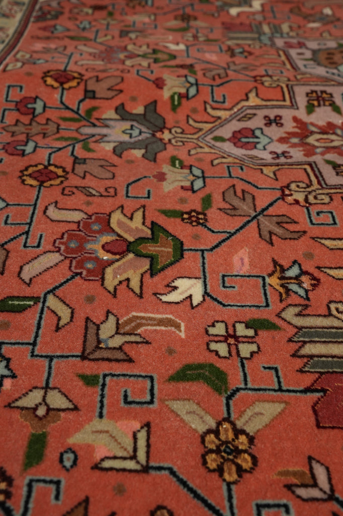 Close-up of a patterned rug with a warm coral-red base and intricate floral and geometric motifs in beige, green, blue, and brown.