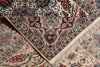 Load image into Gallery viewer, A close-up of an ornate Persian-style area rug with a cream base and intricate floral motifs in blue, red, green, and brown, featuring a folded corner that reveals the detailed pattern and cream fringe.