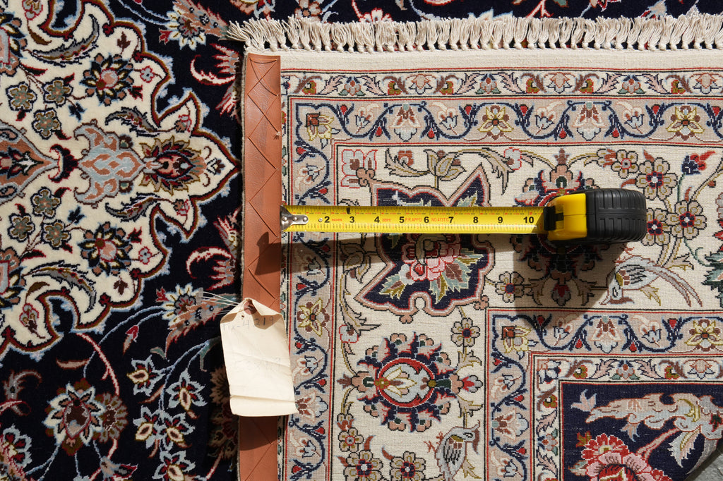 A close-up of an ornate patterned area rug with a yellow tape measure laid horizontally to show the rug's width, alongside a brown leather border and a hanging fabric tag.