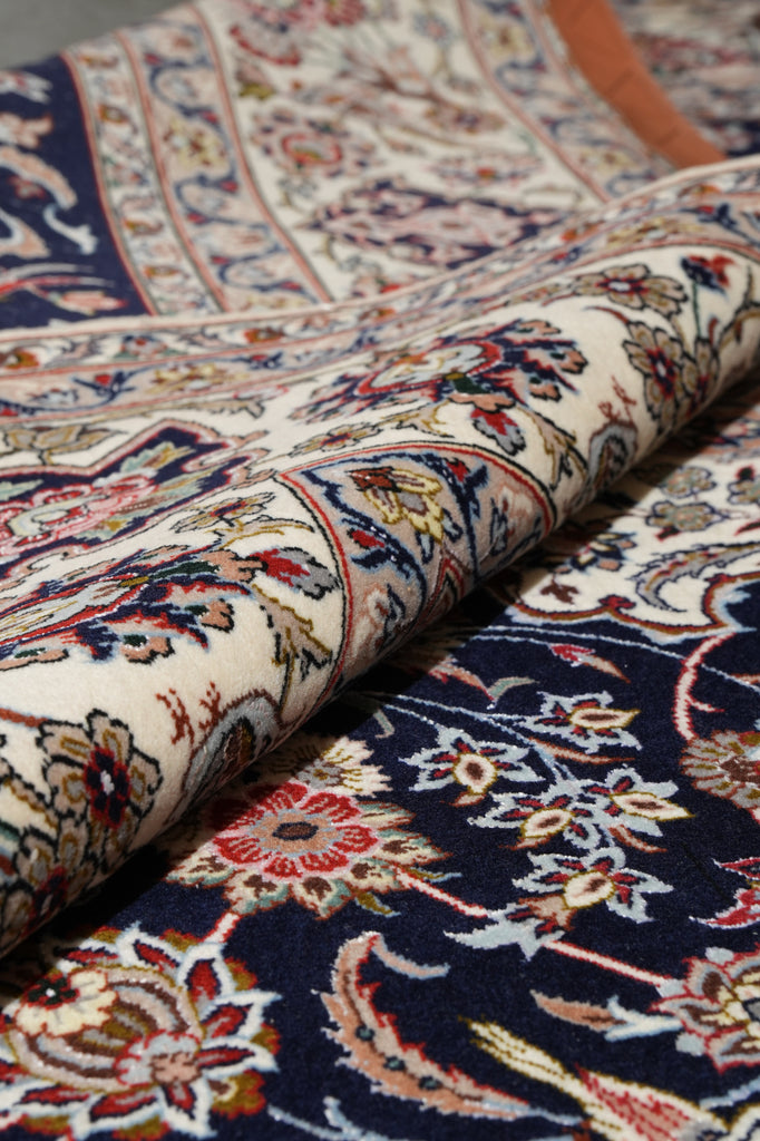 A close-up of a folded traditional oriental rug showing detailed floral and vine patterns in cream, navy blue, red, and beige.