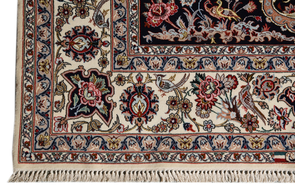 Close-up of an ornate patterned rug corner showing cream ground with navy, red, beige, and gold floral and bird motifs, plus a fringed edge.