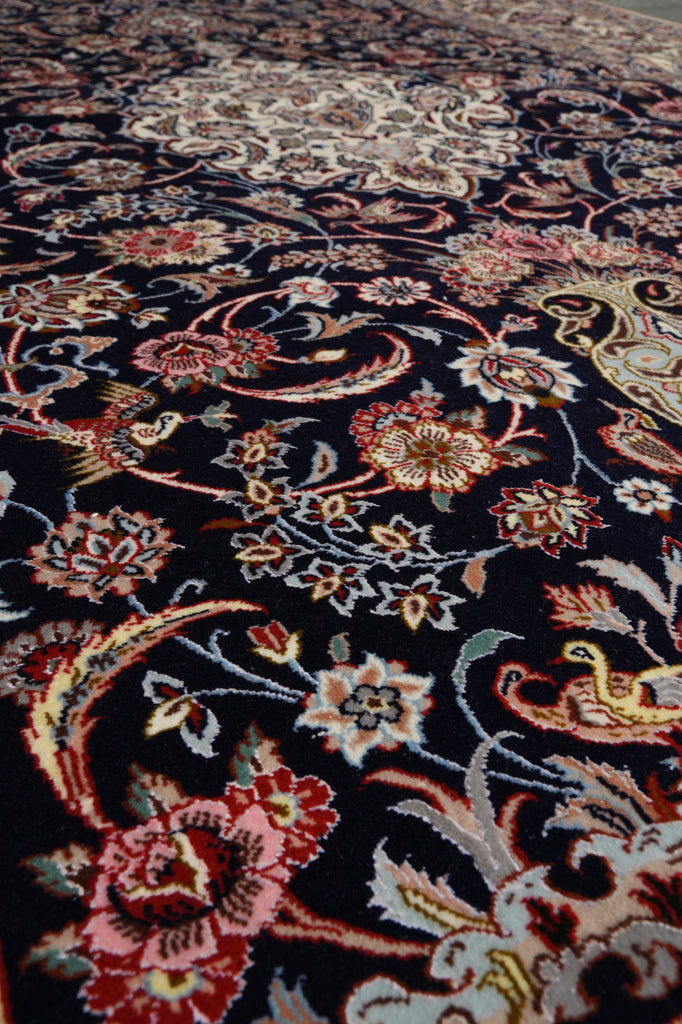 A close-up view of an ornate area rug with a dark navy blue background and intricate floral, paisley, and bird motifs in red, pink, beige, cream, and light blue.