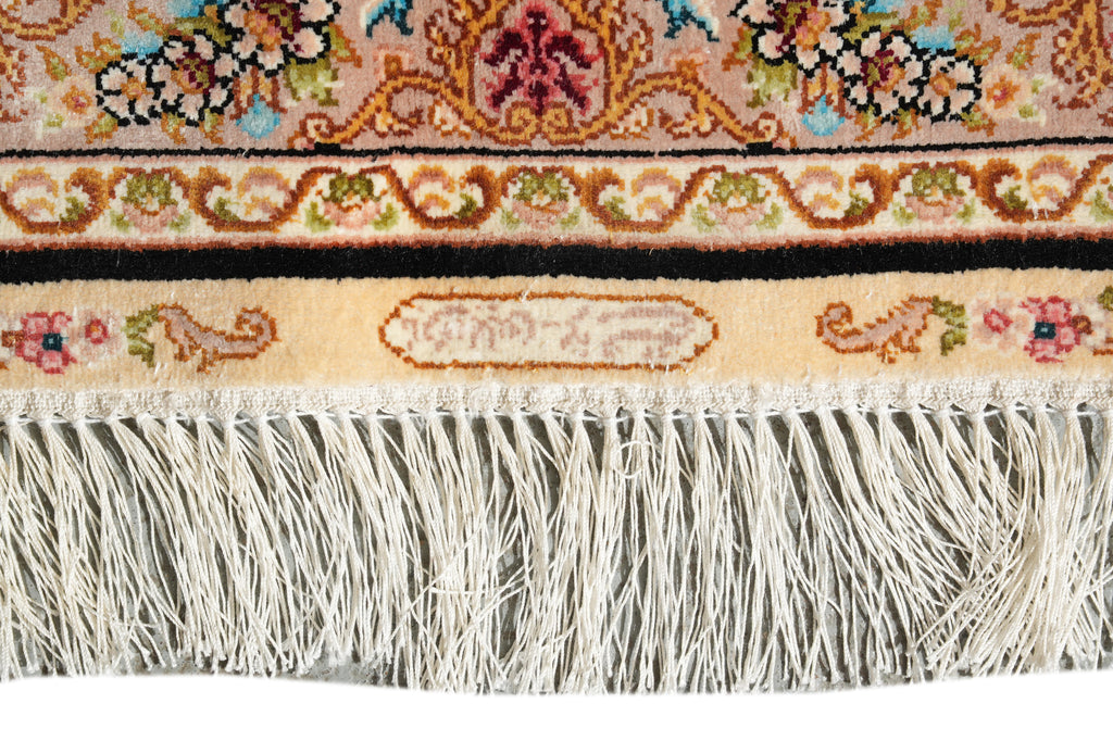 Close-up of a patterned rug showing ornate floral and scroll motifs in beige, brown, black, blue, green, and pink, with a cream fringe along the bottom edge.