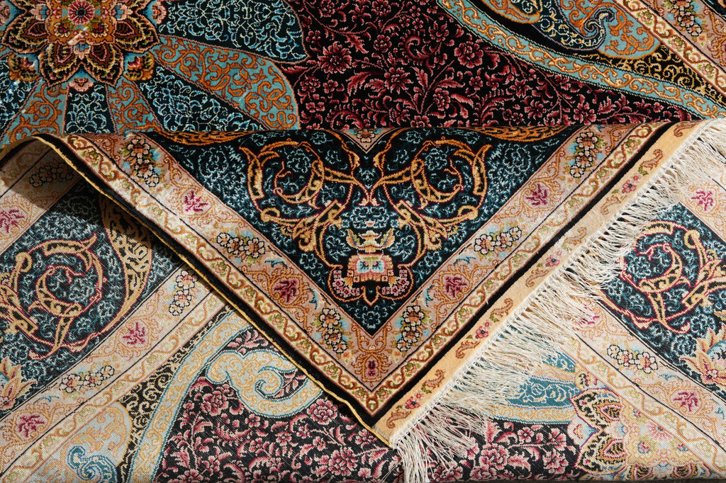 A close-up of an ornate patterned rug with folded sections showing its intricate design, fringed edge, and rich color palette of navy, burgundy, gold, and beige.