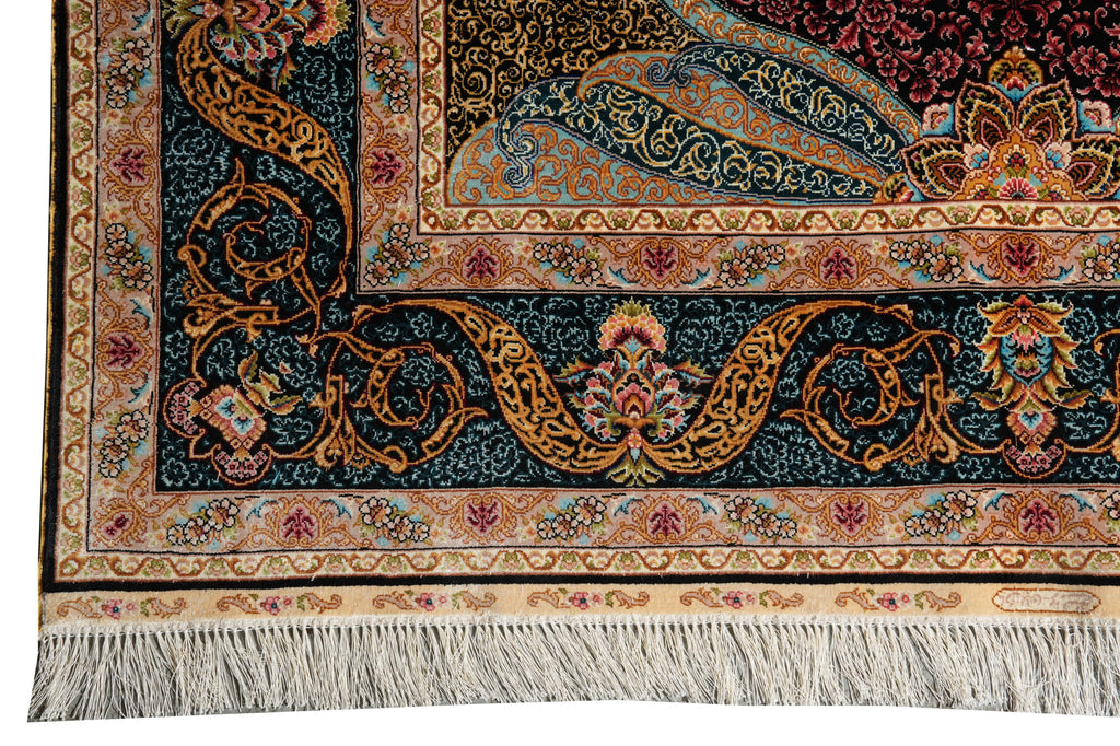 Close-up of an ornate patterned rug corner showing a dark navy field with intricate gold, beige, red, and blue floral motifs, plus a cream fringe along the bottom edge.
