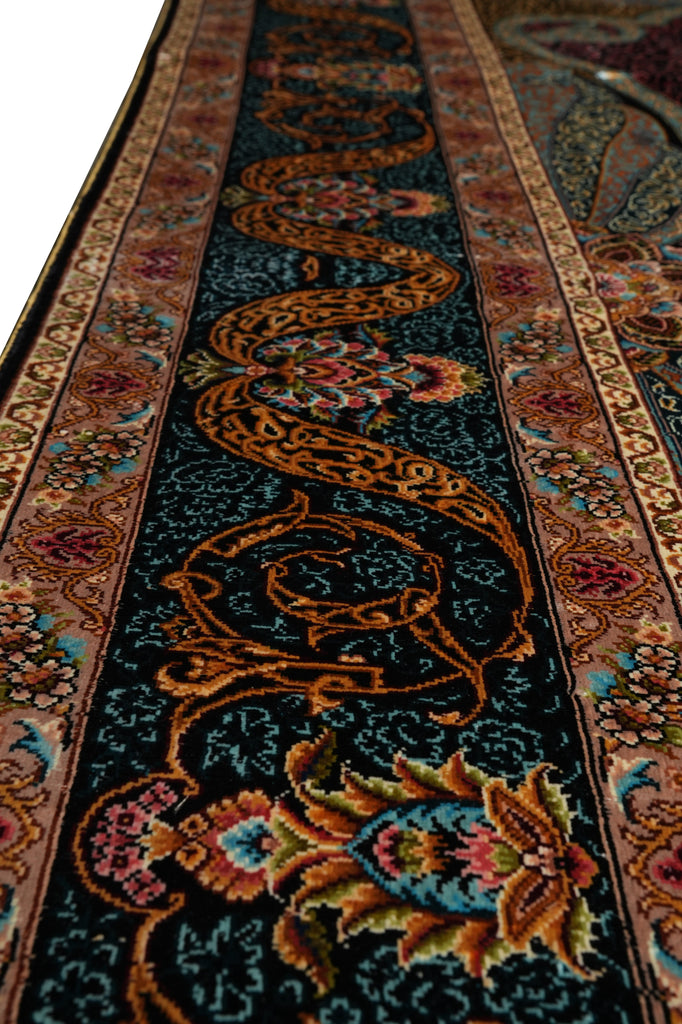 A close-up view of an ornate area rug with a dark navy blue center field, featuring a large golden-brown scrolling motif and colorful floral details in pink, teal, and gold, bordered by patterned bands.
