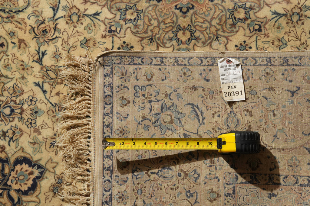 A close-up of a patterned area rug with beige, blue, and brown floral motifs and fringe, shown with a yellow tape measure and a price tag on top.