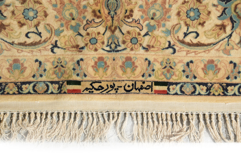 Close-up of a patterned rug with cream, beige, blue, and brown floral motifs, a decorative border, and cream fringe along the bottom edge. Persian text is woven into the border.