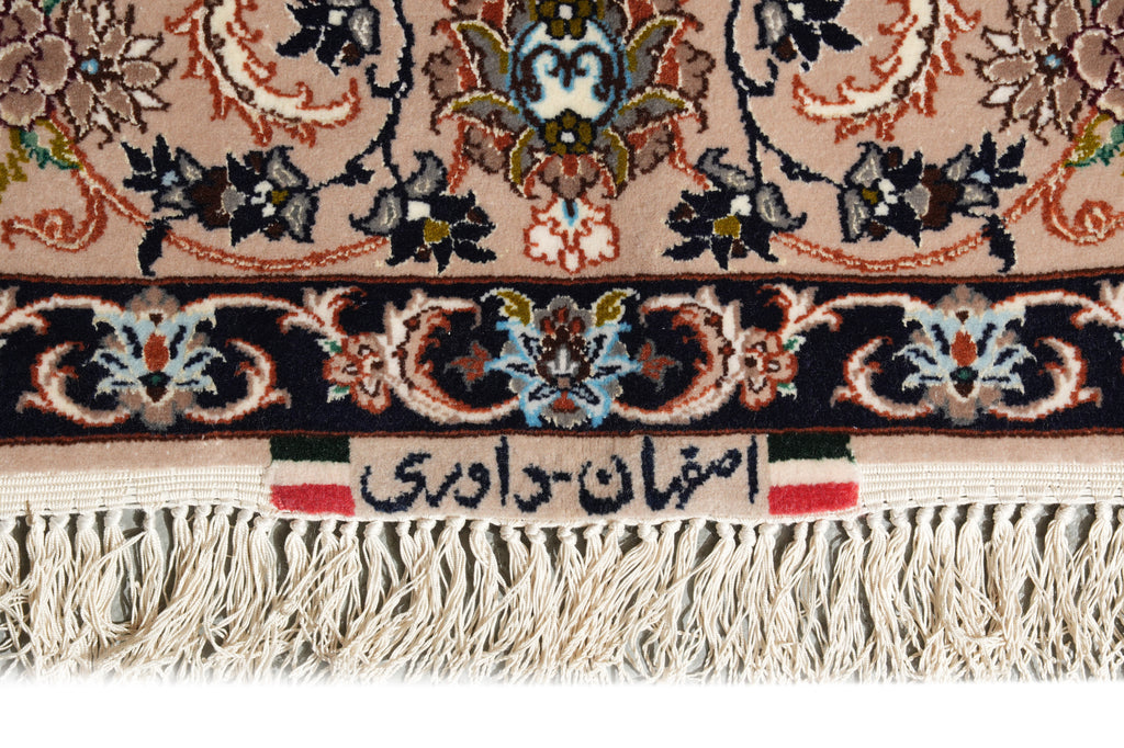 Close-up of a patterned rug with cream fringe, featuring a beige field, a dark navy border with floral motifs, and a central label with Persian text.