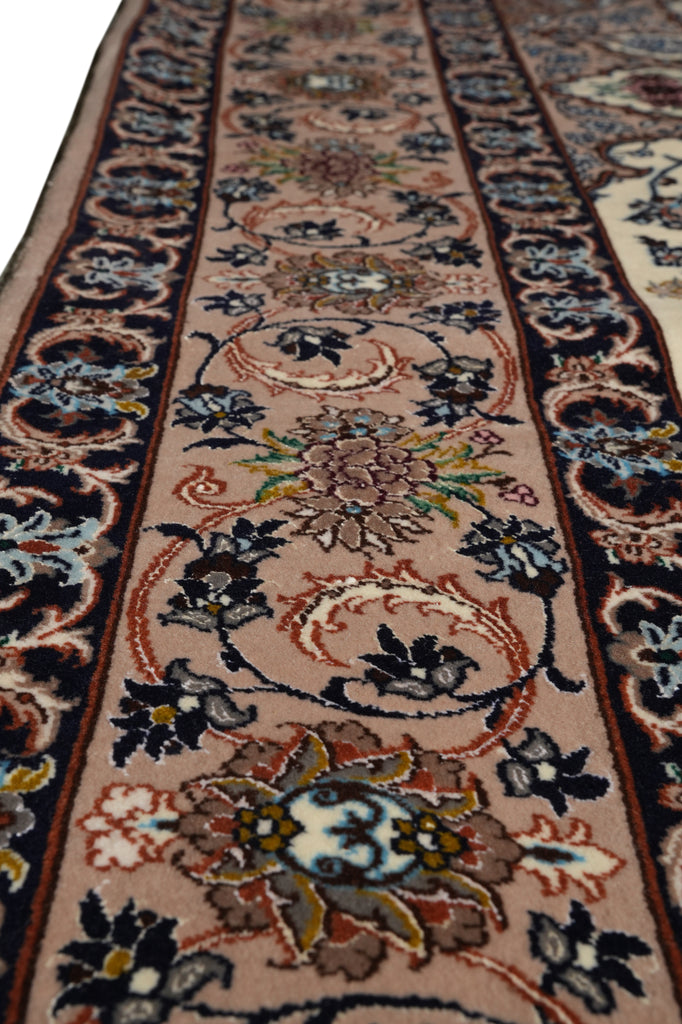 A close-up view of an ornate area rug with a traditional floral pattern. The rug features a central field of dusty pink and beige with dark navy blue borders and intricate floral motifs in blue, green, brown, and cream.