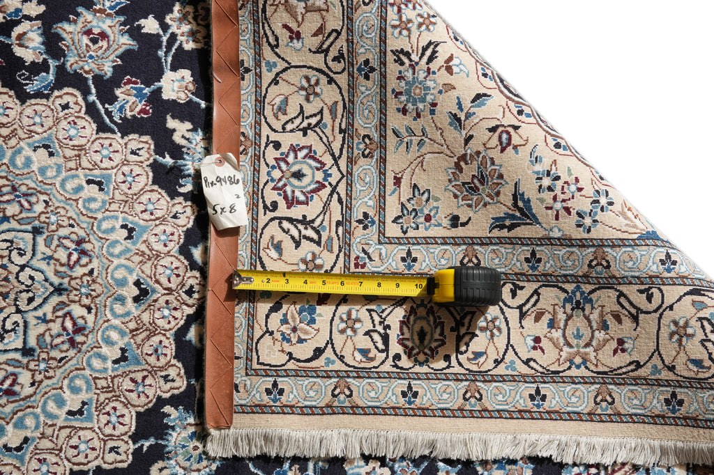 A close-up of a patterned area rug with one corner folded back to show the underside and fringe, alongside a yellow tape measure and a small handwritten size tag on a white background.