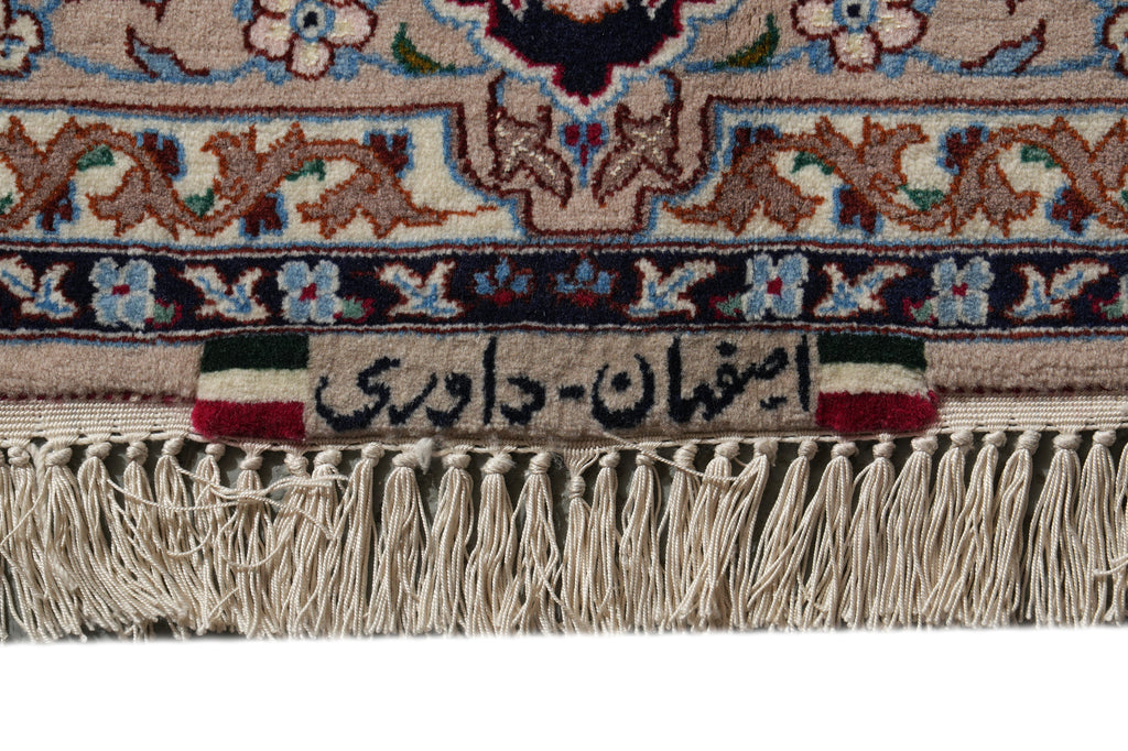 Close-up of a patterned rug with beige, brown, blue, and red woven floral motifs, a fringed bottom edge, and a small woven label with Persian text and green, white, and red side bands.