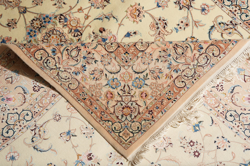 A close-up of a folded ornate area rug showing its intricate floral and scroll pattern in beige, peach, blue, and brown tones on a cream background.