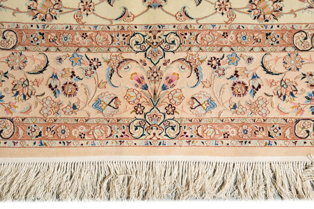 A close-up of an ornate patterned rug showing a cream and beige base with intricate floral motifs in blue, pink, and brown, finished with a thick cream fringe along the bottom edge.