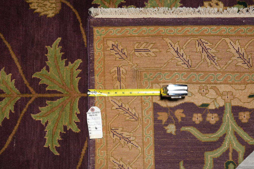 A close-up of an area rug corner with a tape measure laid horizontally to show its dimensions. The rug features a dark plum purple field with green leaf motifs and a tan central border with geometric patterns.