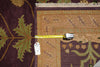 Load image into Gallery viewer, A close-up of an area rug corner with a tape measure laid horizontally to show its dimensions. The rug features a dark plum purple field with green leaf motifs and a tan central border with geometric patterns.