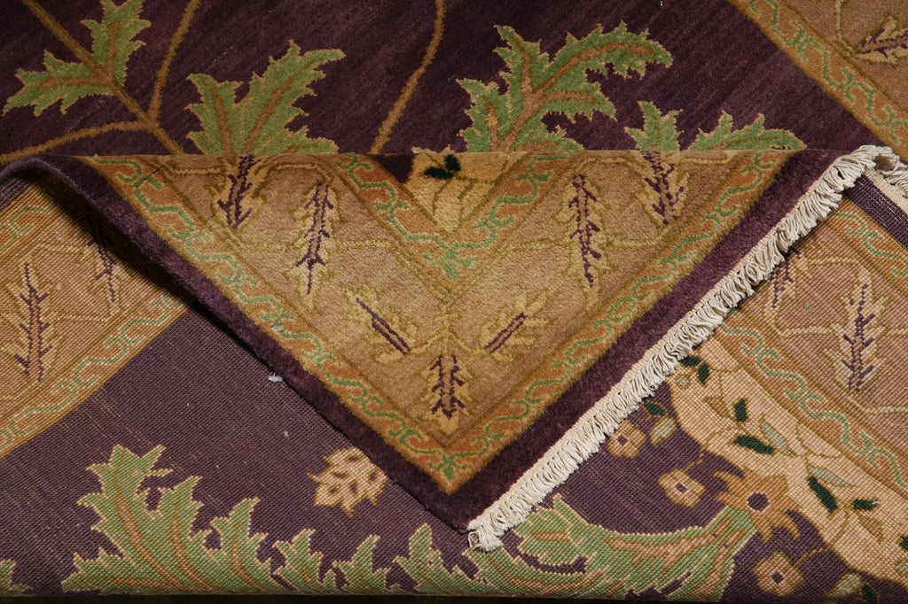 A close-up of an ornate patterned rug with a folded corner showing cream fringe. The design features a deep purple border with green leaf motifs and a central tan section with geometric and floral details.