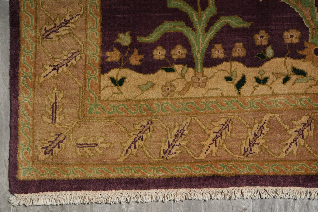 Close-up of a woven rug corner showing a dark plum purple field with a green plant motif, a tan border with leaf patterns, and cream fringe.
