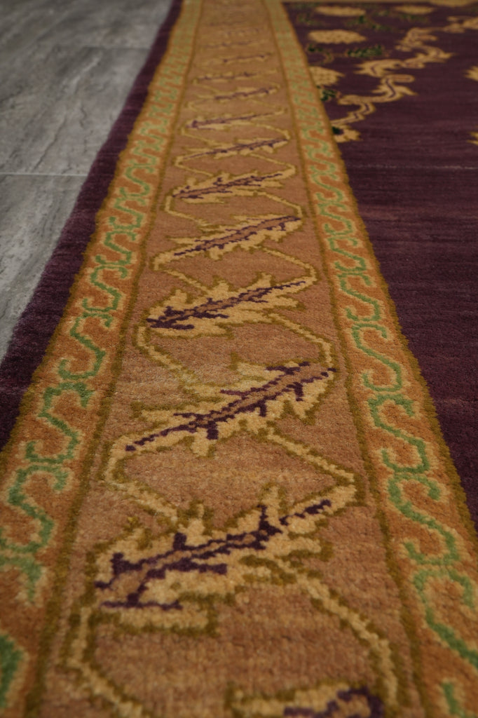 A close-up of a patterned area rug with a central tan and brown field, green and purple geometric motifs, and a dark purple outer border, shown on a gray floor.