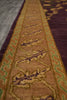 Load image into Gallery viewer, A close-up of a patterned area rug with a central tan and brown field, green and purple geometric motifs, and a dark purple outer border, shown on a gray floor.