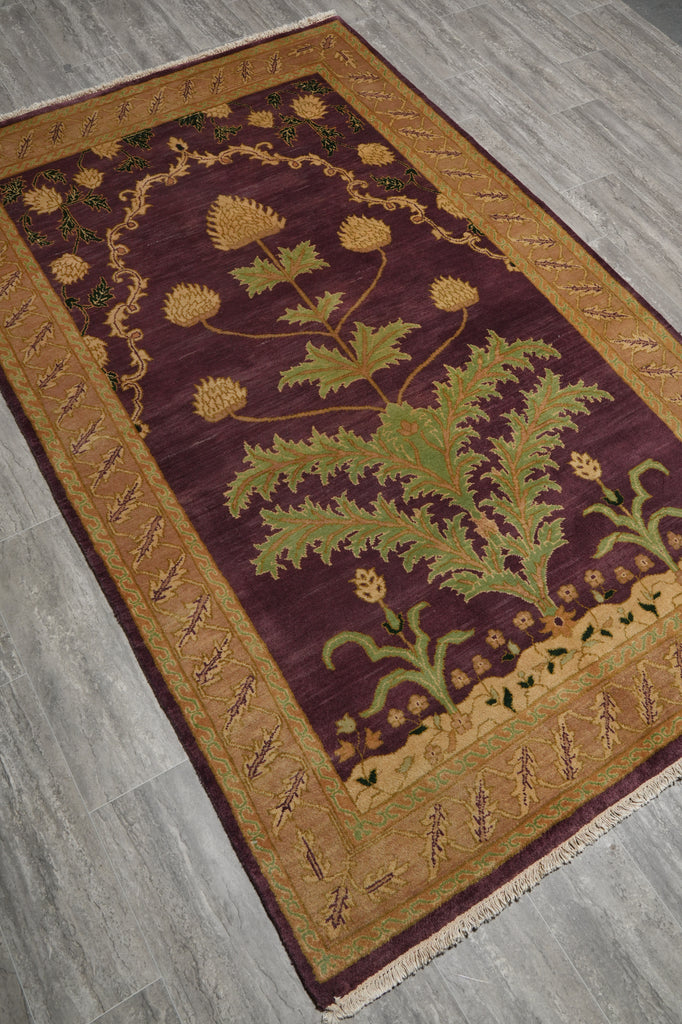 A rectangular area rug with a deep burgundy center and a wide gold border, featuring a large green thistle design in the middle. The rug is shown from an overhead angle on a gray wood-look floor.