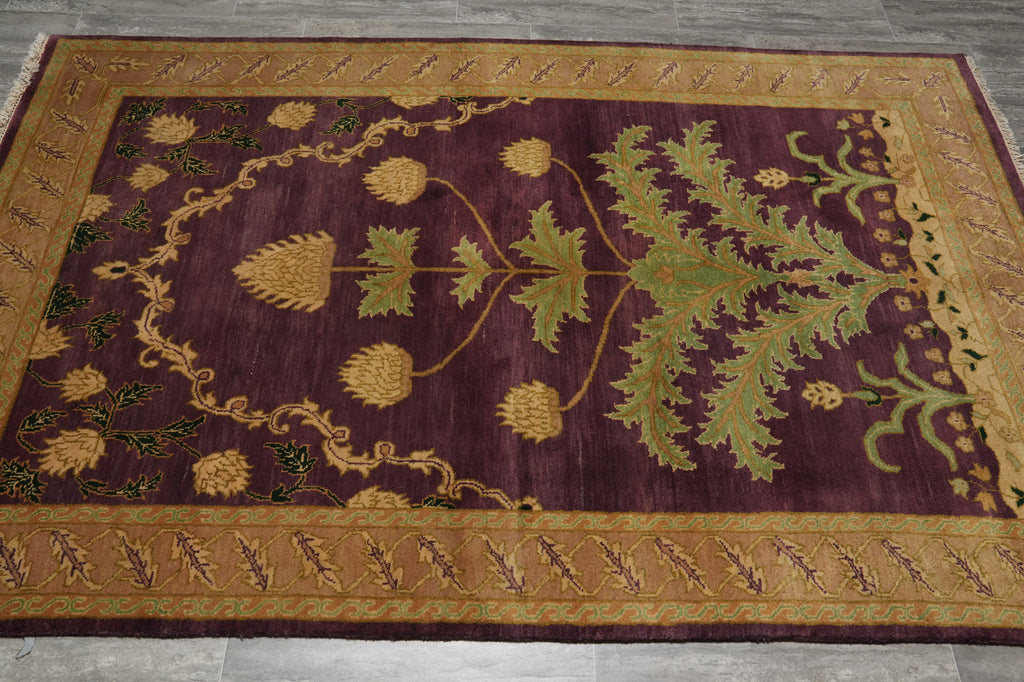 A rectangular area rug with a deep burgundy center field featuring a large green botanical motif and gold thistle-like floral elements, surrounded by a wide gold and beige patterned border, laid flat on a gray tiled floor.