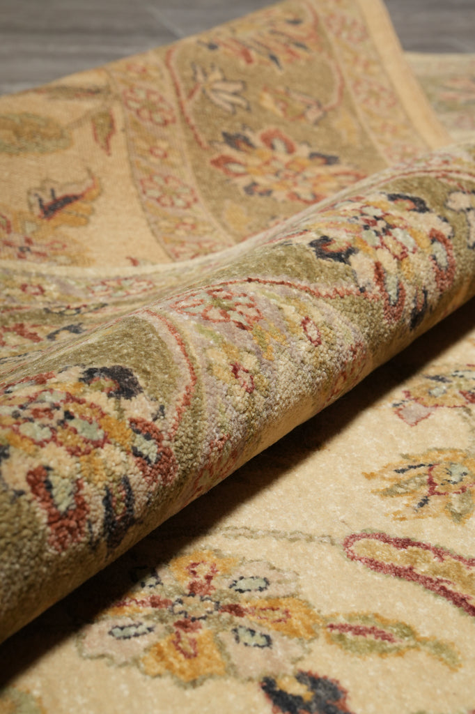 A close-up of a rolled area rug showing its detailed traditional floral and paisley pattern in beige, olive green, rust red, and dark accents.