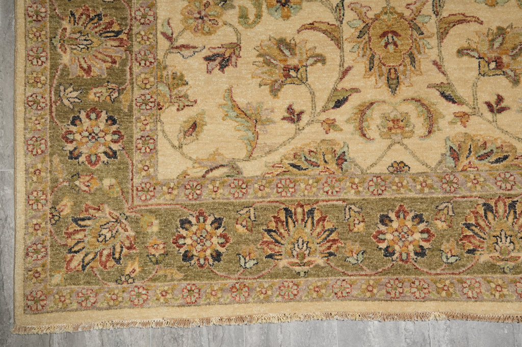A close-up of an ornate traditional rug corner showing a cream field with floral motifs and a wide olive-green border with multicolored floral details and fringe.