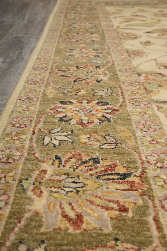 A close-up view of a traditional patterned area rug with a sage green base, featuring floral motifs in beige, rust red, and dark blue, laid out on a gray floor.