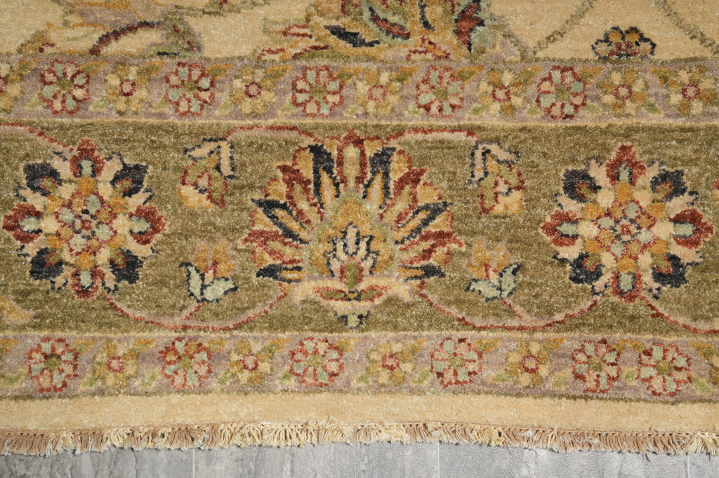 Close-up of a patterned area rug with a central floral medallion and border motifs in muted olive green, beige, rust red, and navy tones, with visible fringe along the bottom edge.