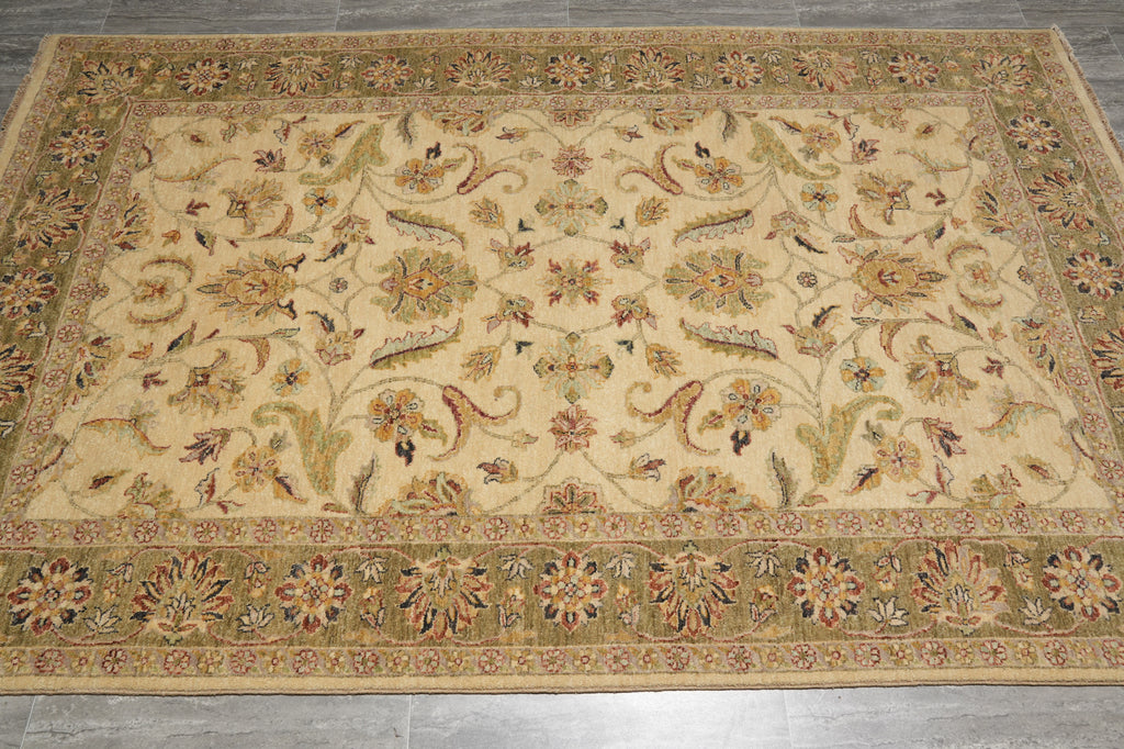 A rectangular area rug with an intricate traditional floral and paisley pattern, featuring a beige center field and a wide olive-green border, laid out on a gray tiled floor.