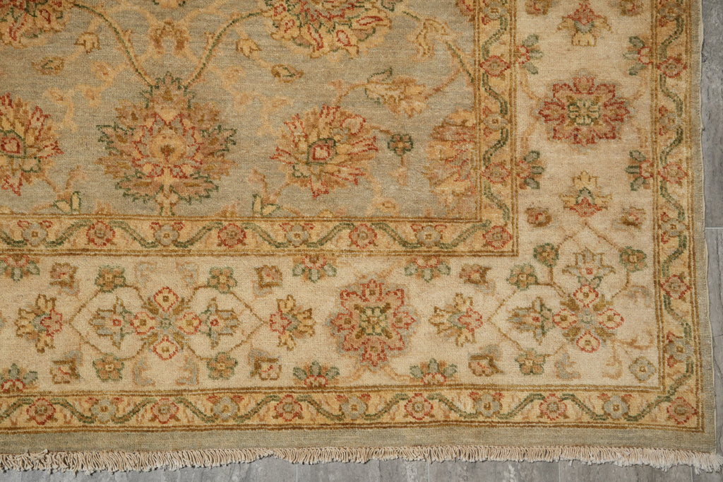 A close-up of an ornate area rug with a traditional floral and medallion pattern. The rug features a warm beige base with intricate motifs in muted green, terracotta red, and soft blue, finished with a fringed edge.