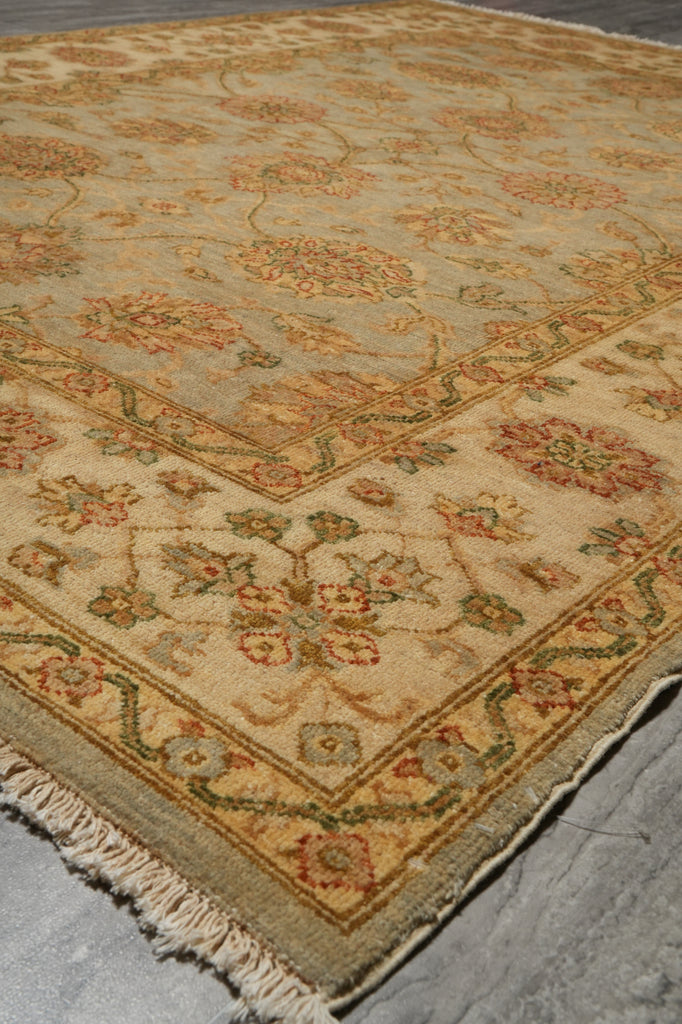 A close-up of a traditional patterned area rug with a beige base, featuring floral motifs in muted orange, green, and red, with a fringed edge on a gray concrete floor.