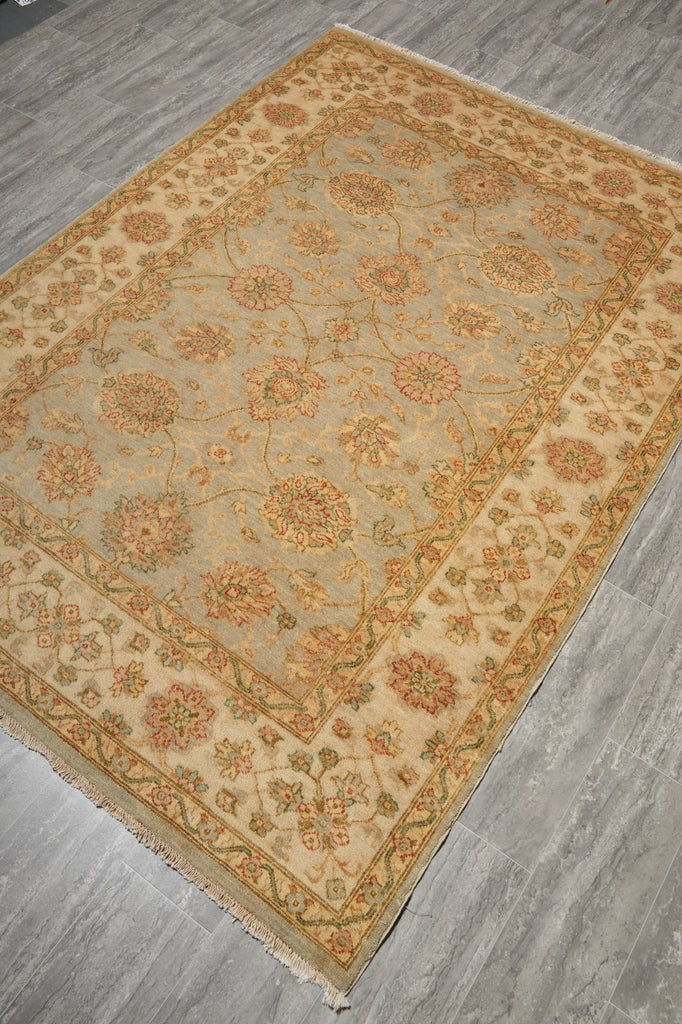 A rectangular area rug with an intricate floral pattern, displayed on a gray wood-look floor. The rug features a beige center field with a warm tan border and motifs in muted orange, green, and brown.