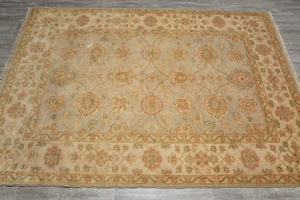 A rectangular area rug with an intricate floral and vine pattern, featuring a beige center, muted green-gray background, and rust-orange borders, laid out on a gray tiled floor.