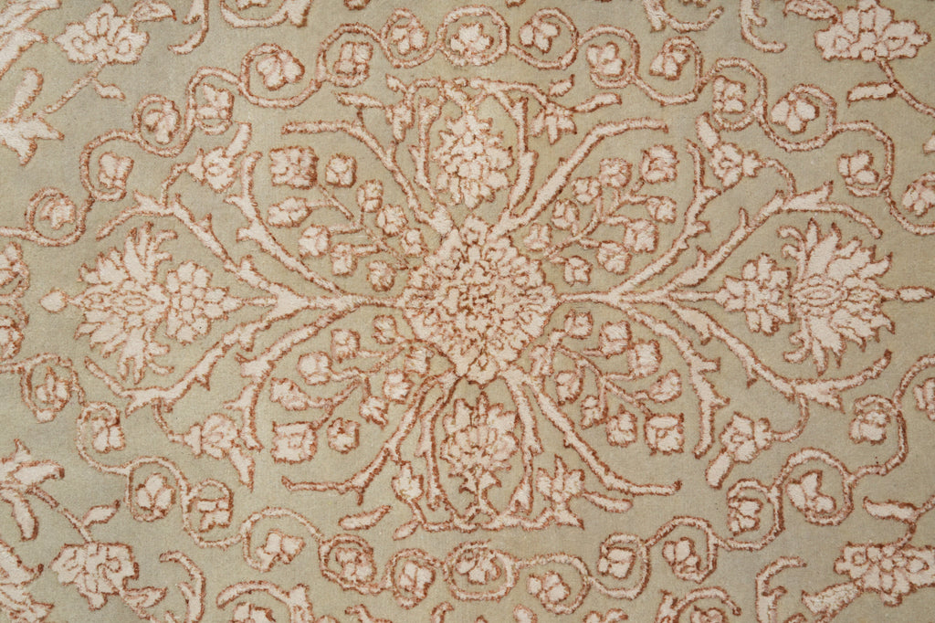A close-up of a textured fabric or rug featuring an ornate, symmetrical floral and vine pattern in a dusty rose or terracotta color over a muted sage green background.