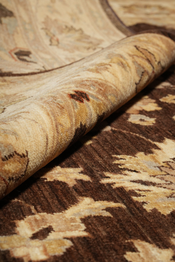 A close-up of a patterned area rug with one corner rolled up to show the pile and texture. The color palette features warm beige, cream, and dark brown tones in an intricate floral or botanical design.