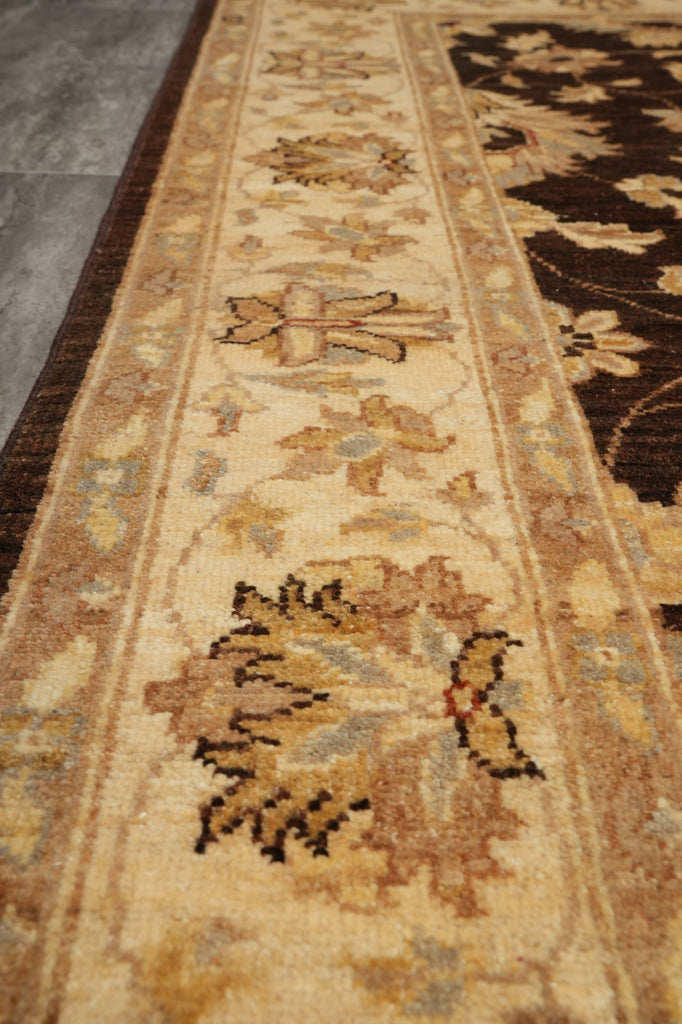 A close-up view of a traditional area rug with a beige center field, featuring floral motifs in brown, gold, and muted blue-gray, bordered by dark brown sections.