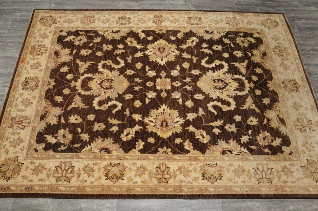 A rectangular area rug with an ornate traditional floral pattern, featuring a dark brown center field and a beige border, laid out on a gray wood floor.