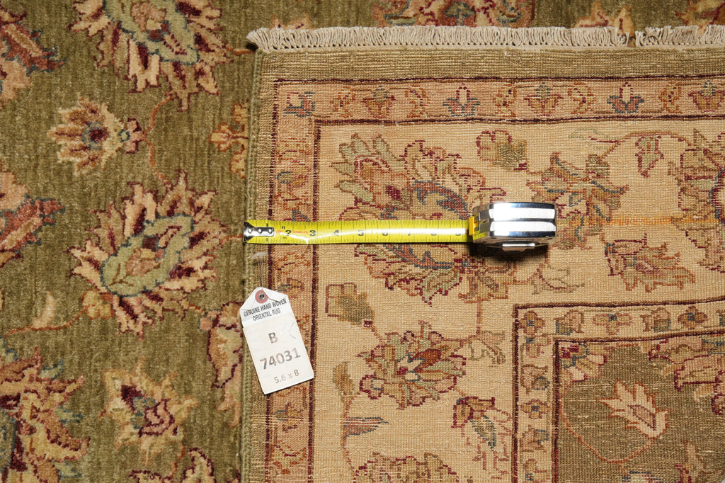 A close-up of a patterned area rug with a tape measure and price tag placed on it to show dimensions. The rug has a beige center with a brown and muted green floral pattern and a fringed edge.