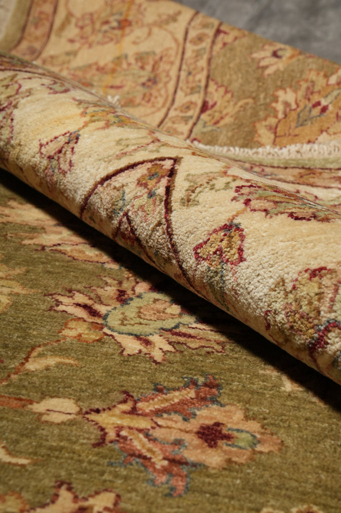 A close-up of a traditional patterned area rug with a rolled section showing its pile and design details. The rug features a warm beige and olive green base with floral motifs in muted red, blue, and yellow accents.